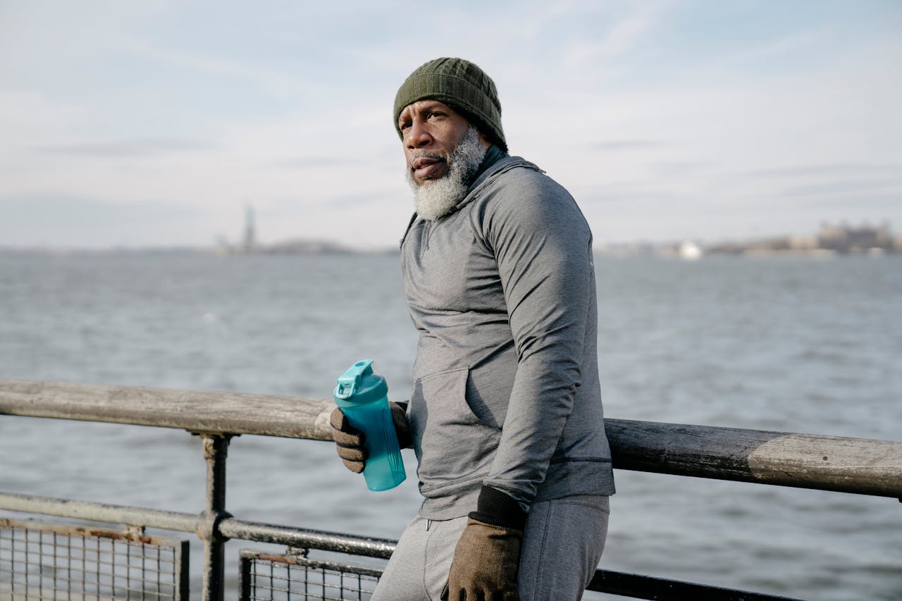 Elderly African American gray haired male wearing warm activewear leaning on metal fence standing near sea with bottle of water and looking away in daytime
